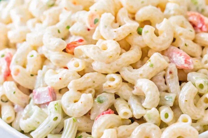 A close-up of a creamy macaroni salad with shrimp, featuring elbow macaroni, diced celery, red bell pepper, and a rich mayonnaise-based dressing, served in a white bowl.
