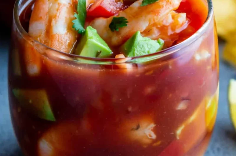 Colorful Mexican shrimp cocktail served in a glass with tomato sauce, avocado chunks, fresh cilantro, and plump shrimp.