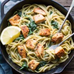 Creamy salmon pasta with tender salmon chunks, linguine, spinach, and a lemon wedge in a black skillet.
