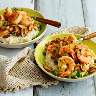 Two bowls of shrimp and grits topped with bacon, mushrooms, and green onions on a rustic wooden table.