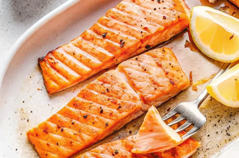 Grilled Coho salmon fillets on a white serving plate with lemon wedges and black pepper seasoning.