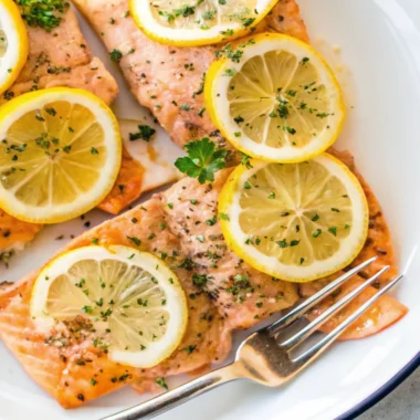 Oven-baked lemon pepper salmon fillets topped with fresh lemon slices and sprinkled with herbs, served on a white plate with a silver fork.