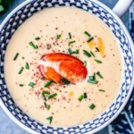 Creamy lobster bisque in a patterned bowl, garnished with a lobster claw, chopped chives, and cracked black pepper.