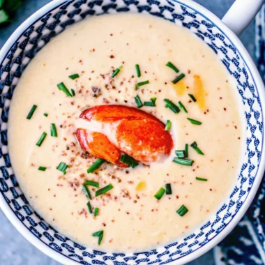 Creamy lobster bisque in a patterned bowl, garnished with a lobster claw, chopped chives, and cracked black pepper.